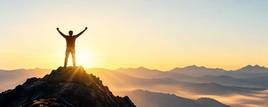 Silhouetted figure standing triumphantly on a mountain peak at sunrise, arms raised, layered misty mountain ranges in the distance