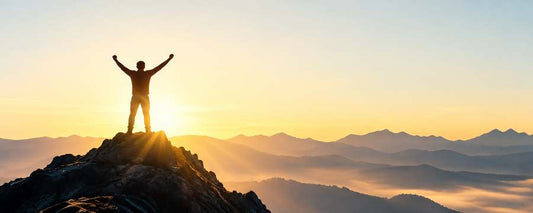 Silhouetted figure standing triumphantly on a mountain peak at sunrise, arms raised, layered misty mountain ranges in the distance